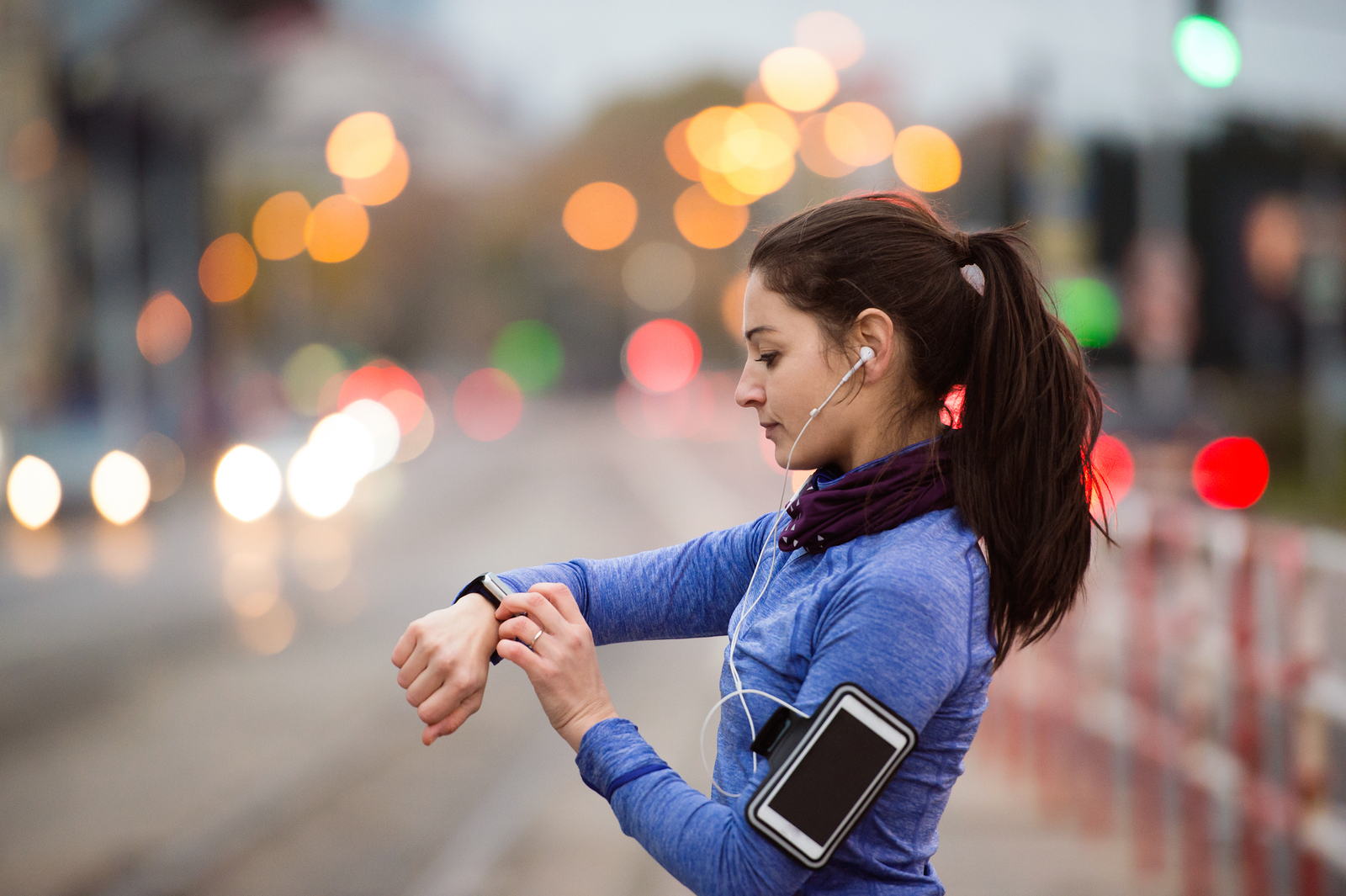 Beautiful young woman in the city with smart phone, smartwatch and earphones, listening music. Using a fitness app for tracking weight loss progress, running goal or summary of her run.